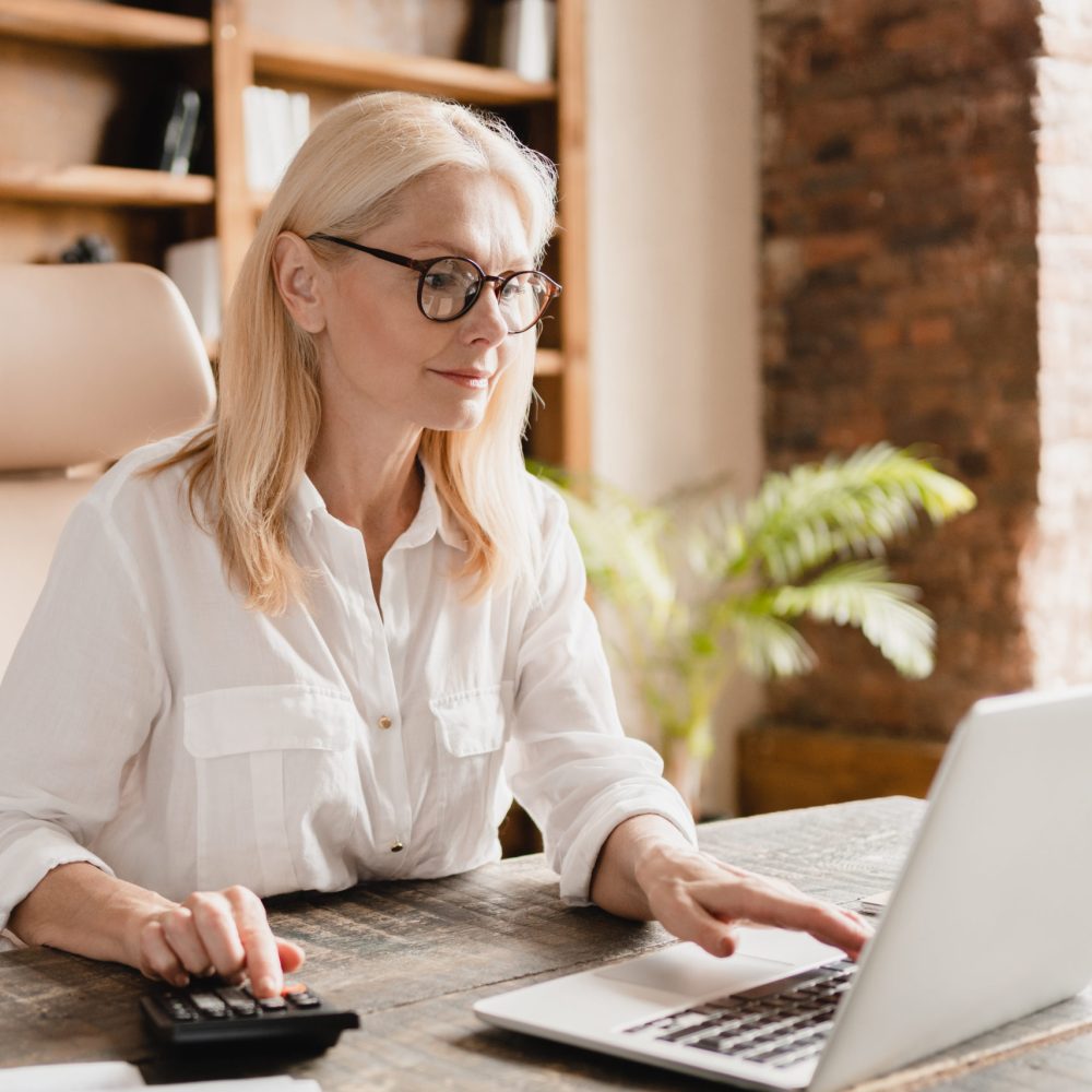 Middle-aged mature businesswoman boss financial advisor counting on calculator funds, money, paying domestic bills, debts, credit loan using laptop in office